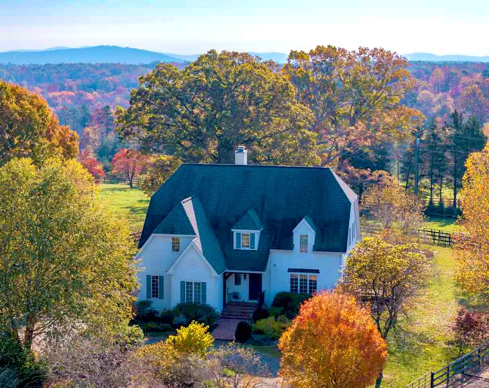 An aerial view of a charming white cottage-style home with a steeply pitched dark roof, nestled among vibrant autumn trees in hues of orange, yellow, and red. The home features dormer windows and a brick walkway leading to the front entrance, surrounded by a landscaped yard. In the background, open fields and rolling hills stretch towards a scenic horizon with distant mountain views, creating a tranquil and picturesque setting.