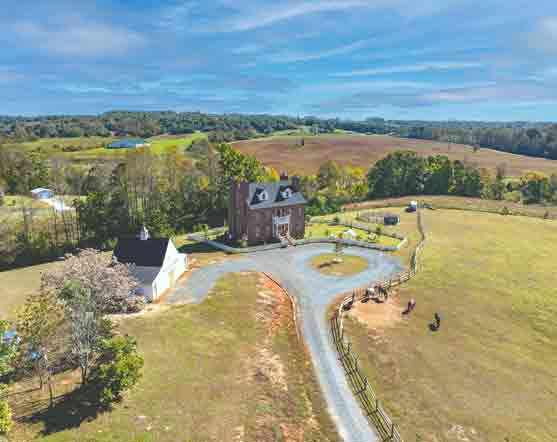 An aerial view of a stately brick manor house set within an expansive pastoral landscape. The home features a circular driveway and is accompanied by a white outbuilding with a dark roof. Surrounding the property are open fields bordered by wooden fencing, with a few horses grazing in the distance. The scene is framed by rolling hills, patches of trees, and a clear blue sky, creating a serene and picturesque countryside setting.