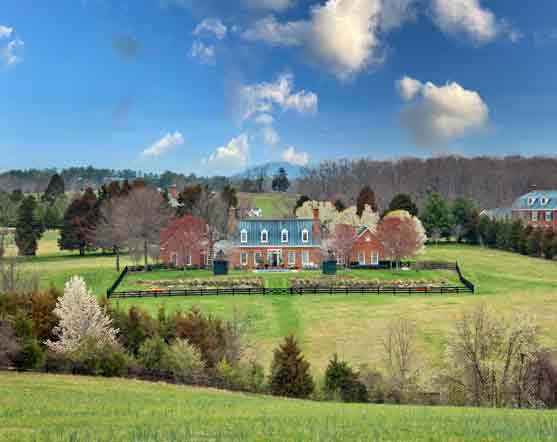 A stunning brick estate surrounded by lush greenery and blossoming trees, featuring a manicured lawn, a black wooden fence, and a backdrop of rolling hills under a vibrant blue sky with scattered clouds.