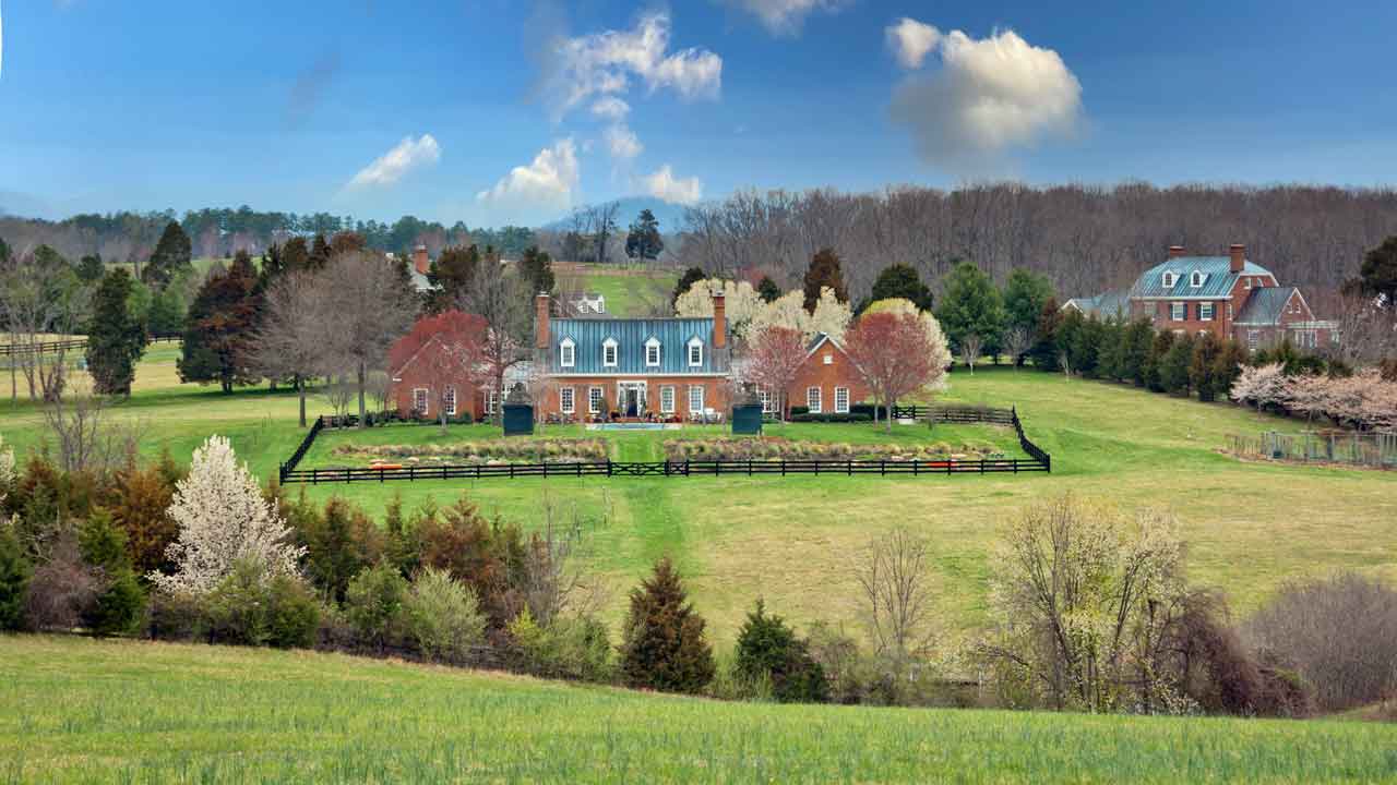 A stunning brick estate surrounded by lush greenery and blossoming trees, featuring a manicured lawn, a black wooden fence, and a backdrop of rolling hills under a vibrant blue sky with scattered clouds.