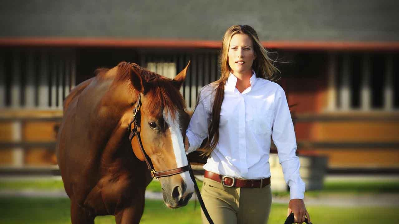 Equestrian woman in a white shirt and khaki pants walking a chestnut horse with a white blaze, set against the backdrop of a red stable on a sunny day.