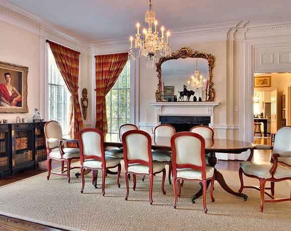 An exquisite dining room showcasing a long polished wooden table encircled by upholstered chairs with ornate wooden frames. A crystal chandelier hangs overhead, casting a warm glow across the space. A gilded mirror above the white marble fireplace reflects the chandelier's light, enhancing the room's elegance. Tall windows draped with red and gold plaid curtains add a classic touch, while a sideboard with books and decorative pieces, along with a framed portrait, completes the refined and traditional ambiance.