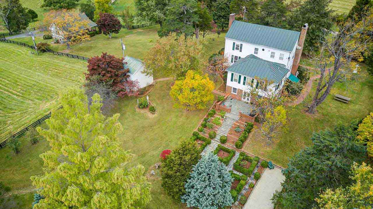 Aerial view of a charming historic home with a green metal roof, landscaped gardens, mature trees, and a detached round-roofed structure. The fenced pasture suggests a perfect country or equestrian setting.