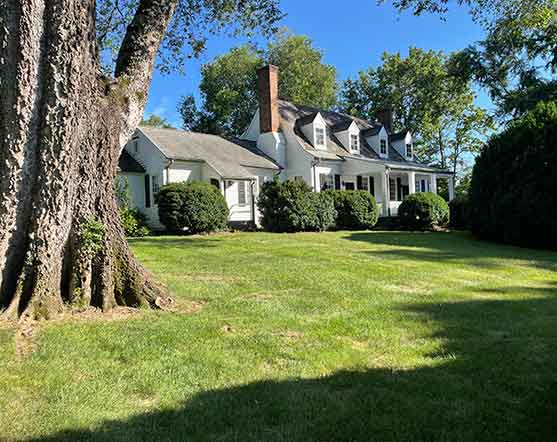 Historic white Colonial-style farmhouse with dormer windows, brick chimneys, and a wide front porch, set on a manicured lawn with mature trees and lush shrubbery in Albemarle County, Virginia.