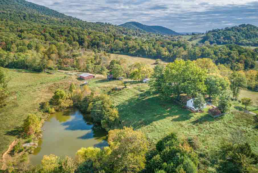 An expansive aerial view of a serene rural property surrounded by lush greenery and rolling hills. The landscape features a tranquil pond reflecting the surrounding trees, a white farmhouse shaded by mature trees, and several outbuildings, including a red barn, scattered across the open fields. The backdrop of wooded hills and distant mountains enhances the peaceful and picturesque setting, ideal for a countryside retreat.